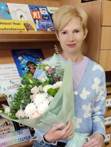 Photo of Maryna Sokolova who has light skin and short blonde hair and is holding a bouquet of flowers while standing in front of a book display.