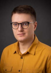 Photo of Oleksii Deikun who has light skin and short dark hair, wearing glasses and an orange button-up shirt.